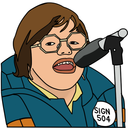 Judy Heumann, known as the mother of disability rights, speaking at the 504 protests in 1977. She has pale skin, short brown hair and glasses, and is speaking into a microphone. She wears a yellow shirt, blue jacket, and a badge that says 'sign 504'.